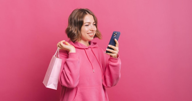 Foto felice giovane donna che tiene la borsa della spesa di carta e utilizza lo smartphone su sfondo rosa