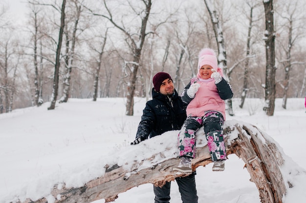 Foto in lontananza di una bambina seduta sul tronco di un albero super eccitata che indossa abiti invernali rosa con il padre in piedi dietro la figlia nella foresta Sfondo sorprendente pieno di colore bianco e neve