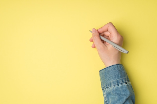 Foto in prima persona della vista dall'alto della mano femminile che tiene una penna grigia su sfondo giallo isolato con copyspace