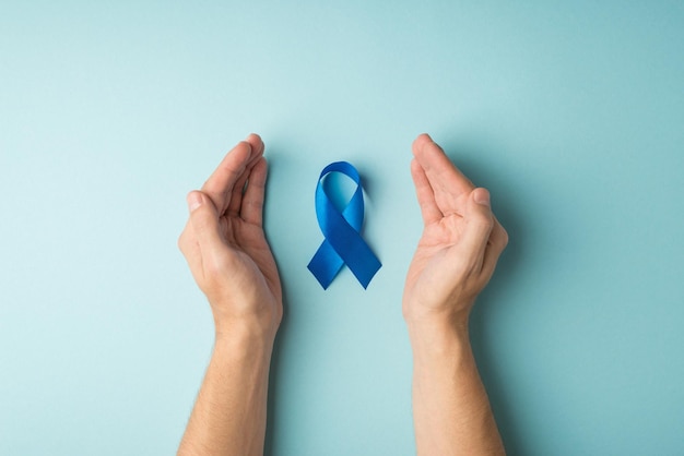 Foto in prima persona vista dall'alto del nastro blu simbolo della consapevolezza del cancro alla prostata tra i palmi maschili su sfondo blu pastello isolato