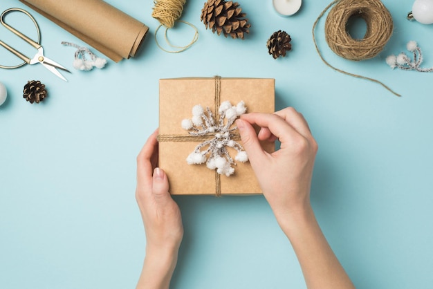 Foto in prima persona vista dall'alto delle mani della donna che decorano la confezione regalo con ramoscello di neve e spago su palle di albero di natale bianche e strumenti artigianali su sfondo blu pastello isolato