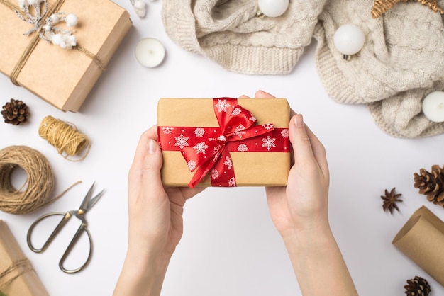 Foto in prima persona vista dall'alto delle mani della donna che tengono la confezione regalo di carta artigianale con fiocco di nastro rosso sopra la sciarpa palle di albero di natale e strumenti artigianali su sfondo bianco isolato