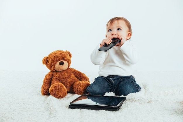 Foto in studio con lo sfondo bianco del viso di un bambino con un cellulare in bocca accanto a un orsacchiotto e un tablet