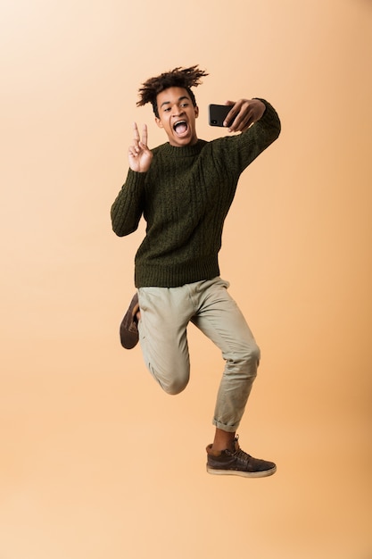 Foto integrale di allegro ragazzo afroamericano che indossa un maglione prendendo selfie sullo smartphone, isolato sopra la parete beige