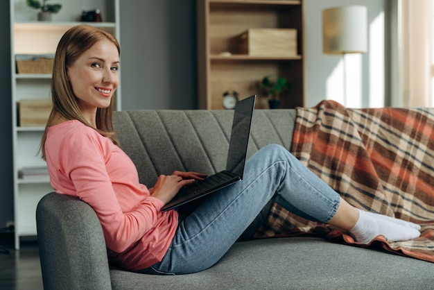 Foto laterale del profilo completo del corpo di una giovane donna con un sorriso positivo felice seduta sul divano e che naviga in un laptop al chiuso