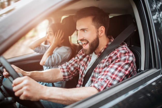 Foto orizzontale del ragazzo emotivo e fiducioso alla guida di un'auto. Sta guardando dritto e urla. La ragazza è stupita e guarda dritto anche sulla strada.