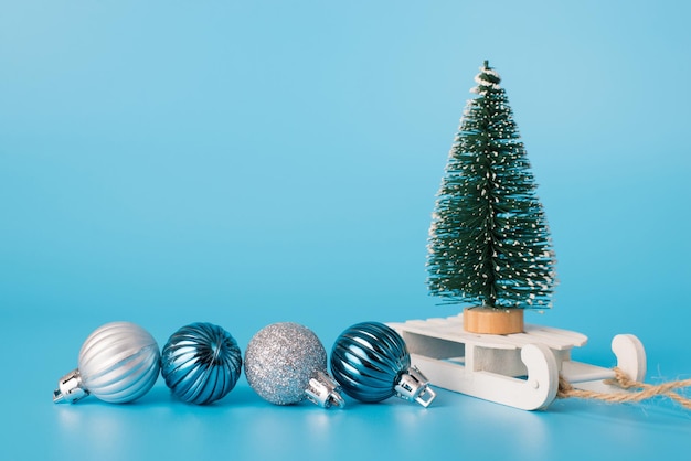 Foto ravvicinata del mini albero di natale in piedi su un po' di slitta in legno bianco con piccole palline accanto isolato su sfondo blu con copyspace