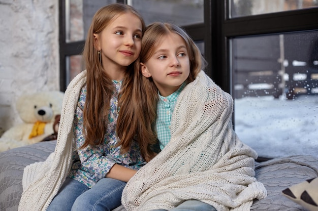 Foto ravvicinata di due bellissime sorelline caucasiche sedute sul davanzale della finestra, coperte da una coperta. Si sono congelati e hanno cercato di riscaldarsi, usando la coperta abbracciandosi l'un l'altro