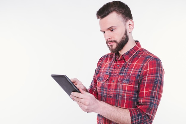 Foto ravvicinata di giovane uomo barbuto felice in camicia a quadri in piedi e utilizzando tablet isolato su sfondo bianco