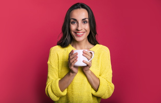 Foto ravvicinata di un'attraente bruna con un maglione giallo, che guarda nella telecamera con un sorriso aperto e tiene in mano una tazza di tè bianca