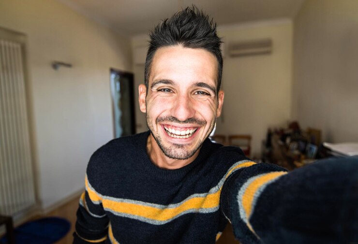 Foto ravvicinata di un bell'uomo hipster che ride in un appartamento - Ragazzo felice si fa selfie a casa - Millennial che sorride alla macchina fotografica - Concetto di stile di vita giovanile