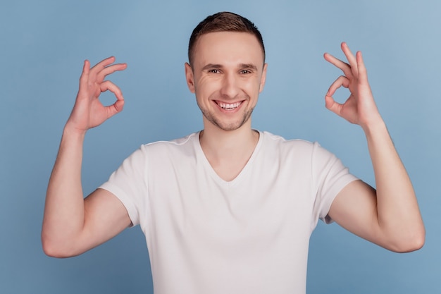 Foto ravvicinata di un ragazzo attraente che si tiene per mano mostrando simboli okey che esprimono accordo sorridente a trentadue denti indossare casual t-shirt rosa isolato sfondo di colore blu