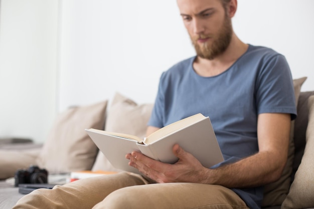 Foto ravvicinata di un uomo seduto sul divano grigio e che legge premurosamente un libro a casa