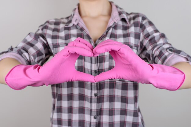Foto ravvicinata di una bella casalinga carina e sicura di sé che fa il cuore con le dita indossando guanti rosa brillante isolati su sfondo grigio