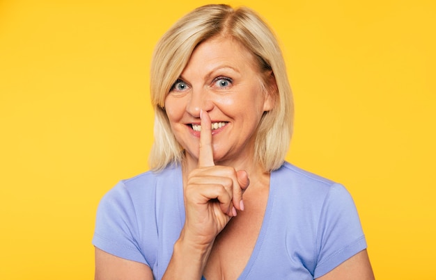 Foto ravvicinata di una bella donna anziana bionda mentre tiene il dito sulle labbra e mostra il gesto di silenzio sul muro giallo