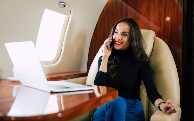 Foto ravvicinata di una bella donna in un abito casual, che sta parlando al telefono e bevendo caffè nero durante il suo volo in jet privato.