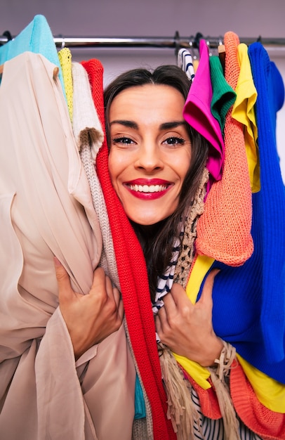 Foto ravvicinata di una bella signora con un grande sorriso, che guarda nell'obiettivo, abbracciando capi di abbigliamento appesi in un armadio, in modo che sia visibile solo il suo viso.