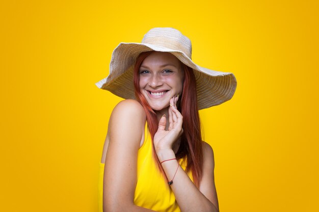 Foto ravvicinata di una donna allo zenzero che sorride alla telecamera su una parete gialla dello studio che indossa un cappello e un vestito