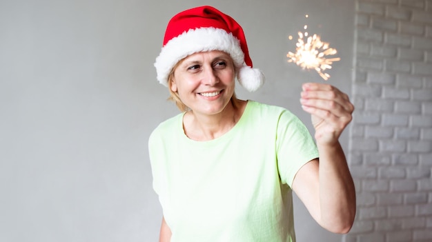 Foto ravvicinata di una donna matura di mezza età eccitata che indossa il cappello di Babbo Natale mentre celebra la festa di Capodanno a casa e si diverte con il fuoco del Bengala in mano