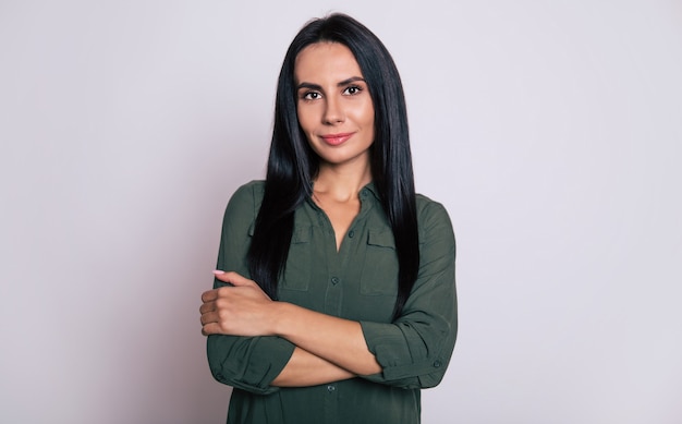 Foto ravvicinata di una giovane ragazza con lunghi capelli nero corvino, che posa con una camicia verde, si abbraccia, guarda nella telecamera con un sorriso rilassato