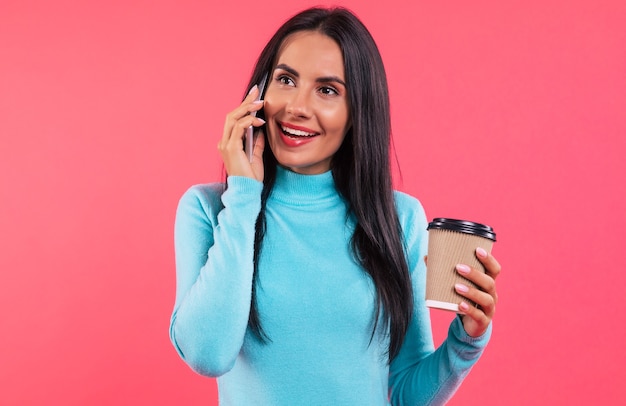 Foto ravvicinata di una splendida donna con un maglione a collo alto blu, che guarda oltre la telecamera e ride mentre parla al telefono e tiene il caffè per andare