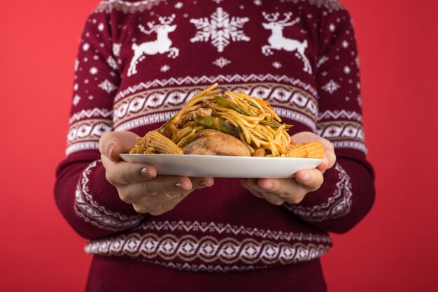 Foto ritagliata del primo piano di una donna in maglione di Natale rosso e bianco che tiene un grande piatto di cibo su sfondo rosso isolato