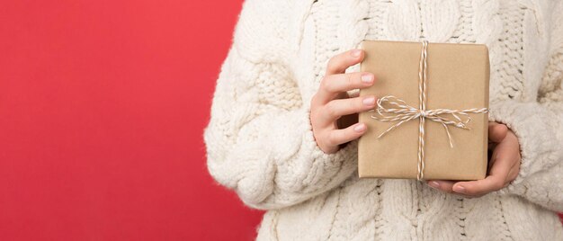 Foto ritagliata del primo piano di una giovane donna in maglione bianco lavorato a maglia che tiene una confezione regalo di carta artigianale con fiocco di spago su sfondo rosso isolato