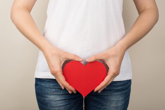 Foto ritagliata del primo piano di una ragazza in maglietta bianca e jeans che tiene un cuore di carta rossa vicino al cavallo su uno sfondo grigio isolato