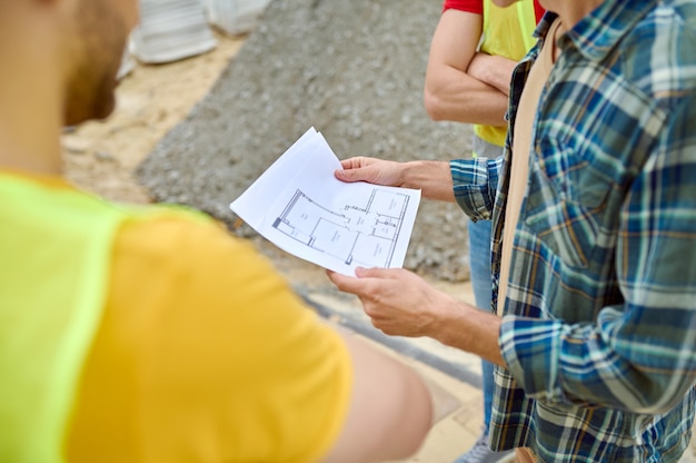 Foto ritagliata di un architetto professionista con progetti di casa nelle sue mani in piedi accanto a due costruttori in giubbotti di sicurezza