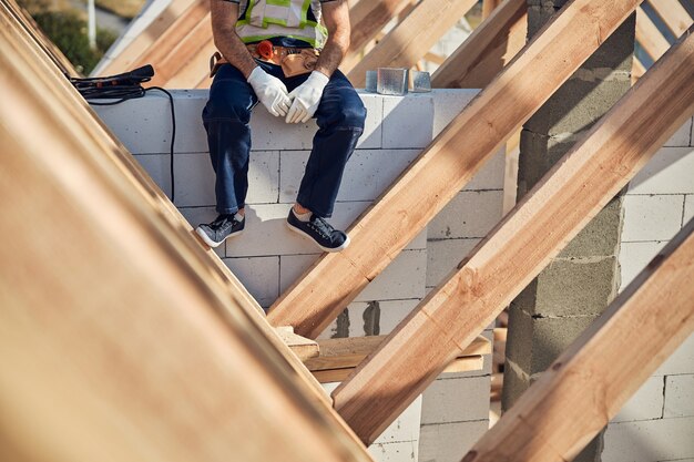 Foto ritagliata di un costruttore stanco seduto su un muro di mattoni di un cantiere e facendo una pausa