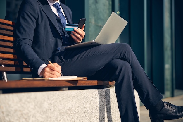 Foto ritagliata di un uomo seduto su una panchina con un laptop e che controlla la carta di credito durante la scrittura