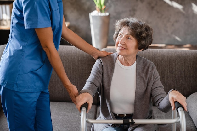 Foto ritagliata di una felice nonna pensionata, un paziente anziano con un telaio per camminare con l'assistenza di un infermiere che si prende cura di lui a casa, che cammina dopo un infortunio traumatico