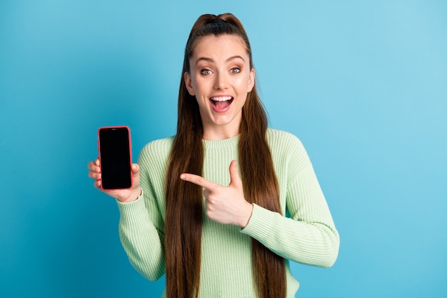 Foto ritratto di ragazza bruna che punta il dito al telefono con spazio vuoto bocca aperta isolata su sfondo colorato blu pastello