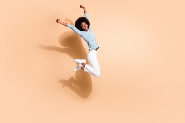 Foto ritratto di ragazza dalla pelle nera che salta in alto due pugni in aria esultando urlando isolato su sfondo color beige pastello