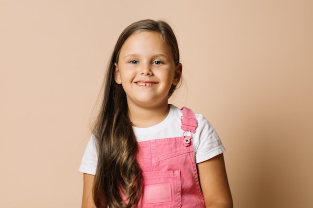 Foto ritratto di una bambina con occhi luminosi e luminosi e un ampio sorriso calmo e felice con i denti che guardano la fotocamera che indossa una tuta rosa brillante e una maglietta bianca su sfondo beige