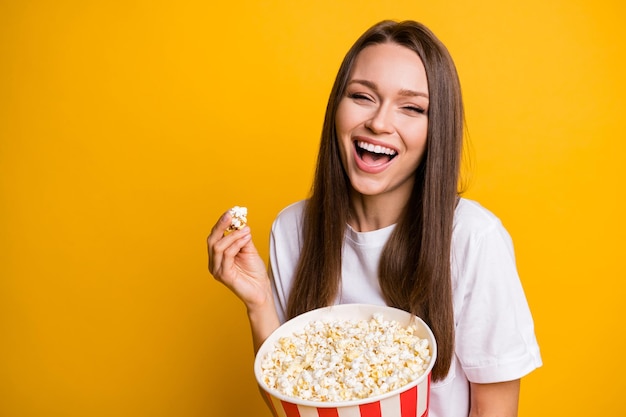 Foto ritratto di una ragazza che ride divertente che tiene in mano una scatola di cartone di pop corn che guarda un film comico al cinema isolato sfondo di colore giallo brillante