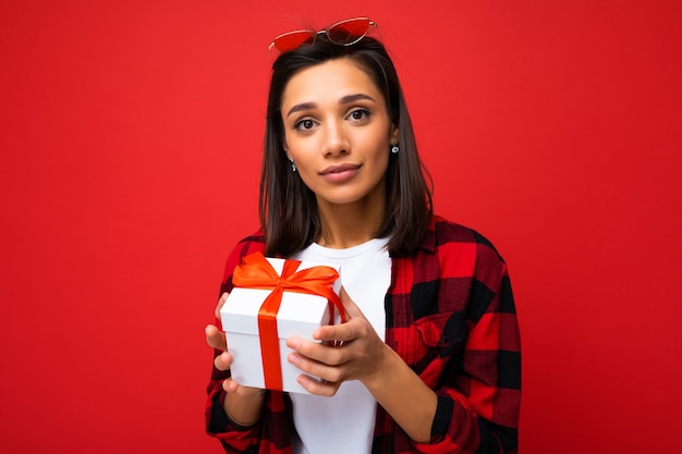 Foto scatto di una giovane donna bruna piuttosto positiva isolata su un muro di sfondo rosso che indossa una t-shirt bianca casual e una camicia rossa e nera che tiene in mano una confezione regalo bianca con un nastro rosso e che guarda l'obbiettivo.