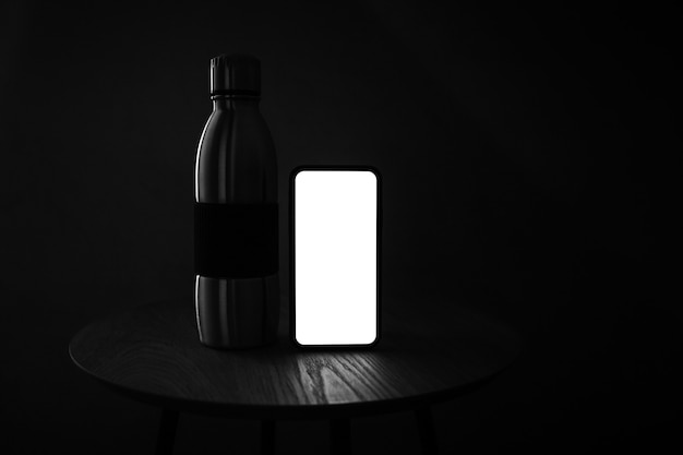 Foto scura in bianco e nero di smartphone con schermo vuoto e bottiglia termica riutilizzabile in acciaio sul tavolo di legno