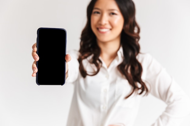 Foto sfocata della donna asiatica sorridente con capelli scuri lunghi che stanno e che mostrano smartphone