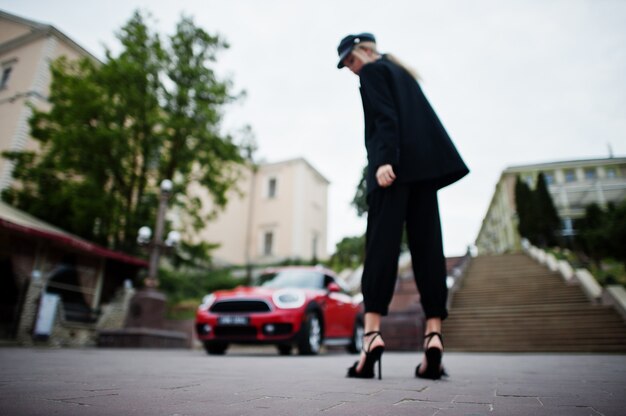 Foto sfocata di bella modella bionda sexy moda posteriore in berretto e in tutto nero con trucco luminoso vicino a city car rossa.