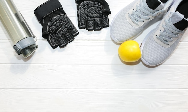 Foto sportiva Stile di vita sano Bottiglia per acqua guanti in pelle e mela Scarpe da ginnastica alla moda Sfondo in legno bianco Perfetta disposizione piatta