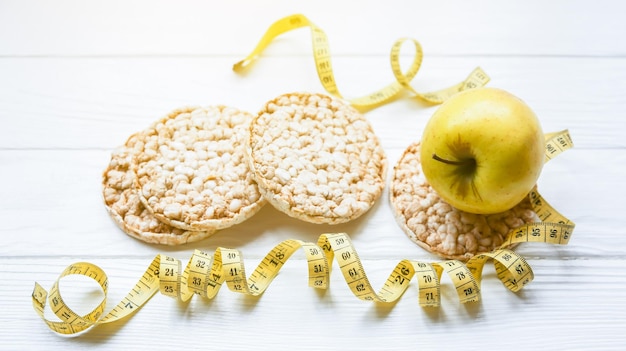 Foto sportiva Stile di vita sano Concetto di fitness Pane e frutta croccanti a basso contenuto di grassi Metro a nastro per corpo in forma Sfondo in legno bianco Perfetto piatto