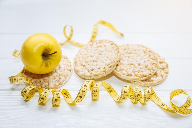 Foto sportiva Stile di vita sano Concetto di fitness Pane e frutta croccanti a basso contenuto di grassi Metro a nastro per corpo in forma Sfondo in legno bianco Perfetto piatto