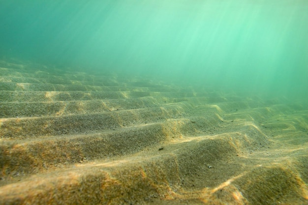 Foto subacquea, piccole "dune" di sabbia riprese in diagonale in modo che in questa prospettiva formino scale, raggi solari provenienti dalla superficie del mare. Sfondo marino astratto.