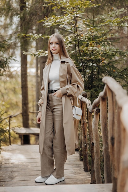 Foto verticale Bella giovane adolescente spensierata con cappotto marrone e jeans bianchi e maglione bianco Ritratto di una bella ragazza con un impermeabile marrone sullo sfondo della foresta e della natura