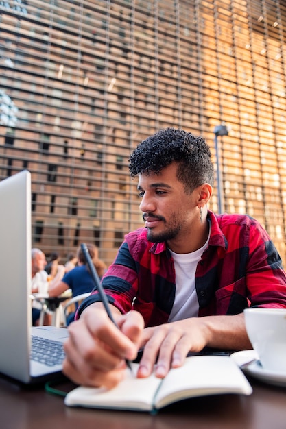 Foto verticale di un giovane latino che studia con il suo computer e prende appunti sulla terrazza di una caffetteria in città, concetto di tecnologia e stile di vita urbano