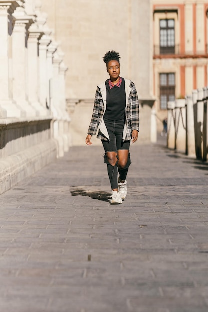 Foto verticale di una giovane donna afro in abiti moderni che cammina per strada in una giornata di sole