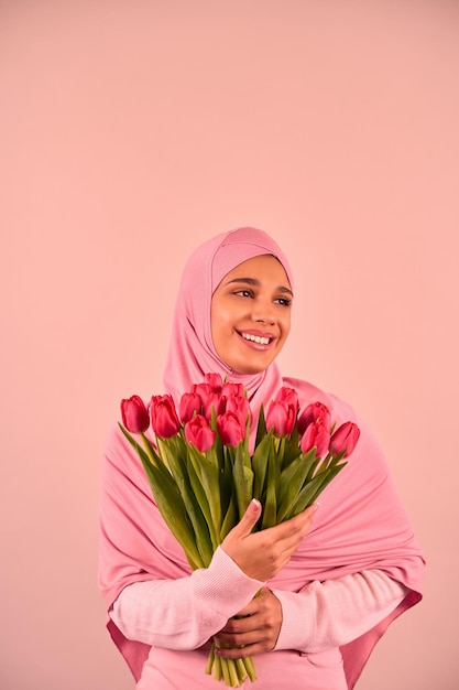 Foto verticale di una giovane donna musulmana in un hijab rosa che tiene un mazzo di tulipani su uno sfondo beige