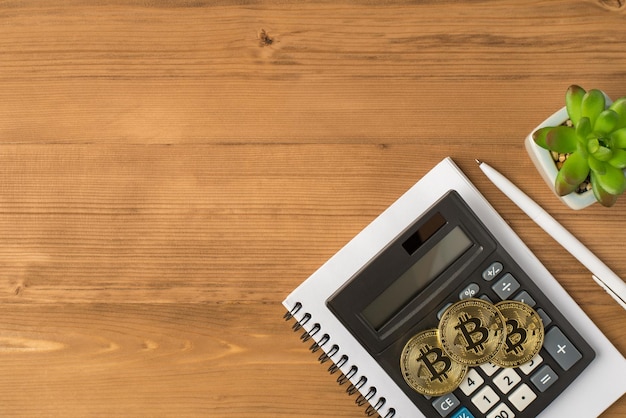 Foto vista dall'alto del posto di lavoro con pianta di penna tre monete d'oro con simbolo bitcoin e calcolatrice su notebook su sfondo tavolo in legno isolato con copyspace