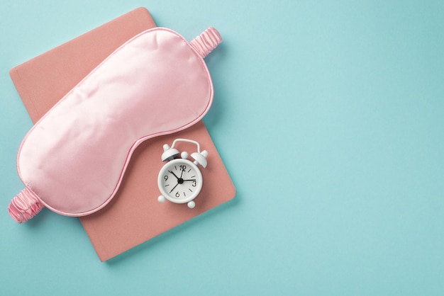 Foto vista dall'alto del taccuino rosa con maschera per dormire sotto di esso e piccolo orologio bianco sul taccuino isolato in un copyspace di sfondo azzurro